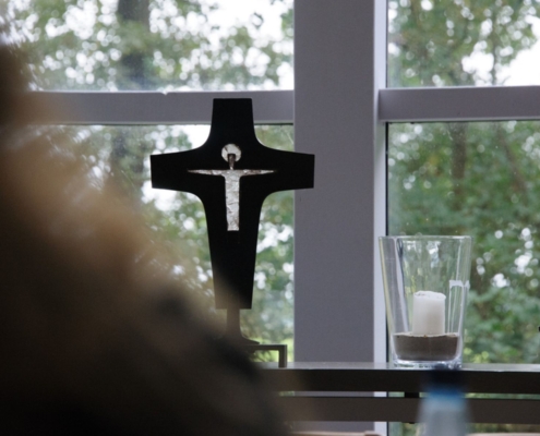 Schwarzes Metallkreuz mit einer stilisierten Christusfigur in der Kapelle des Evangelischen Jugendhofs Sachsenhain in Verden. Im Hintergrund große Fenster mit Blick ins Grüne. Auf dem Altar stehen zwei Kerzen, eine in einem Glasgefäß mit Sand.